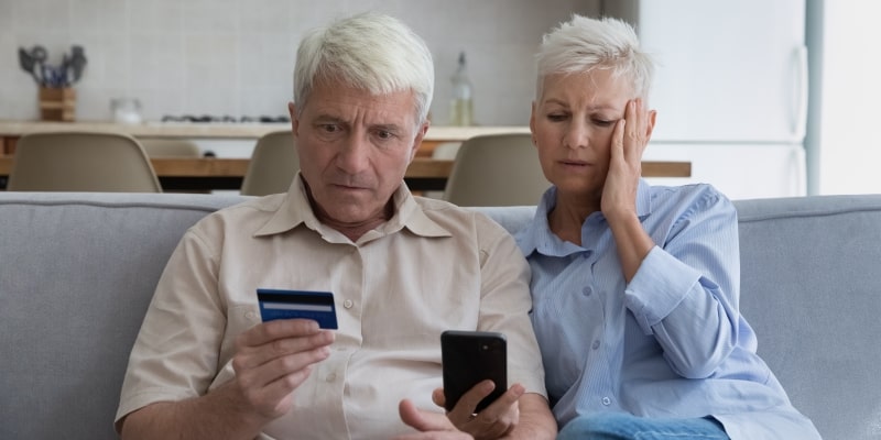 Um casal de idosos supresos de forma negativa. A mulher segura um celular na mão e o homem segura um cartão bancário. Imagem para ilustrar a matéria sobre golpes e fraudes.