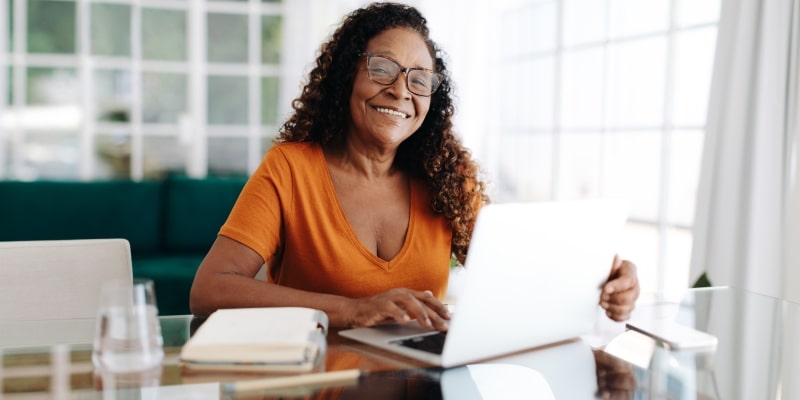 Um mulher idosa e negra, sentada em um escritório e mexendo no computador enquanto sorri. Imagem para ilustrar a matéria sobre como melhorar a saúde mental.