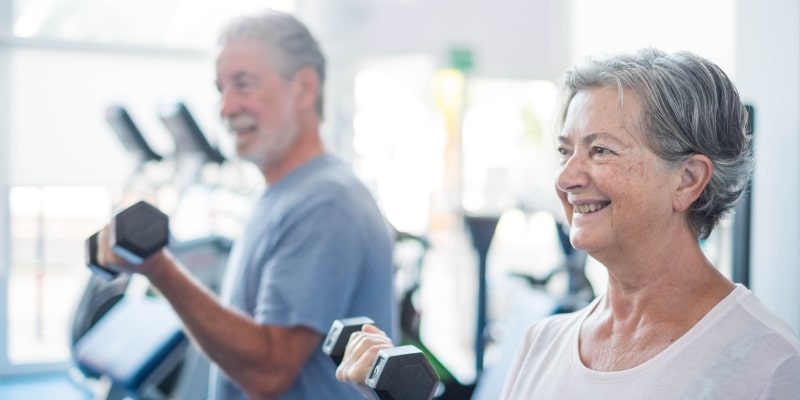 Um casal de idosos levantando alteres em uma academia, fazendo musculação. Imagem para ilustrar a matéria sobre manter a longevidade.