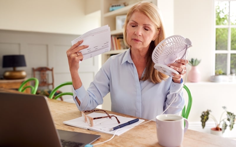 Uma mulher com sintomas de menopausa. Ela está se abanando e usando ventilador enquanto trabalha em casa, na frente de um computador. Imagem para ilustrar a matéria sobre menopausa tabu.