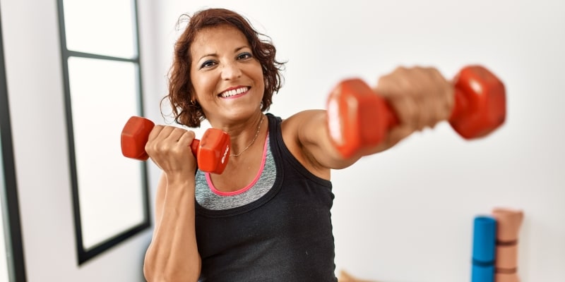 Uma mulher em torno de 50 anos levantando alteres enquanto sorri. Imagem para ilustrar a matéria sobre perda muscular.