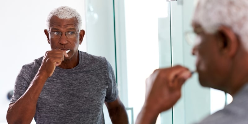 Um homem idoso negro escovando os dentes na frente do espelho. Imagem para ilustrar a matéria sobre saúde bucal de idosos.