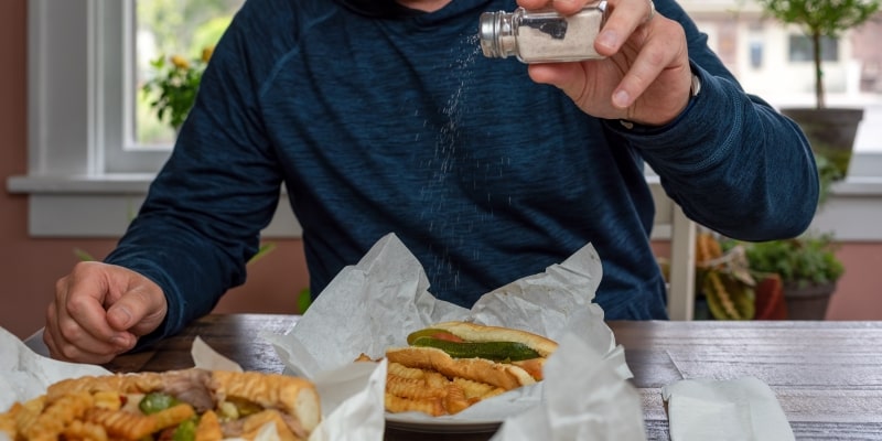 Um homem jogando sal em sanduíches com batata frita. Imagem para ilustrar a matéria sobre alimentos que devem ser evitados.