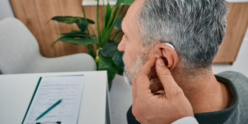 Um senhor, em um consultório médico, enquanto um médico coloca aparelhos auditivos em sua orelha.