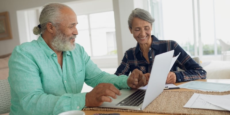 Um casal de idosos sorrindo enquanto vê a Cartilha de Crédito Consciente no notebook e organizam as suas finanças.