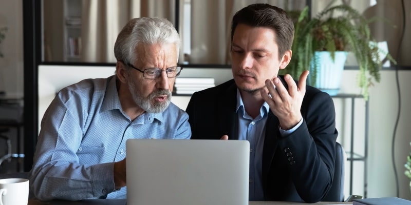 Um homem idoso discutindo com um homem jovem, na frente de um computador. Imagem para ilustrar a matéria sobre etarismo econômico.