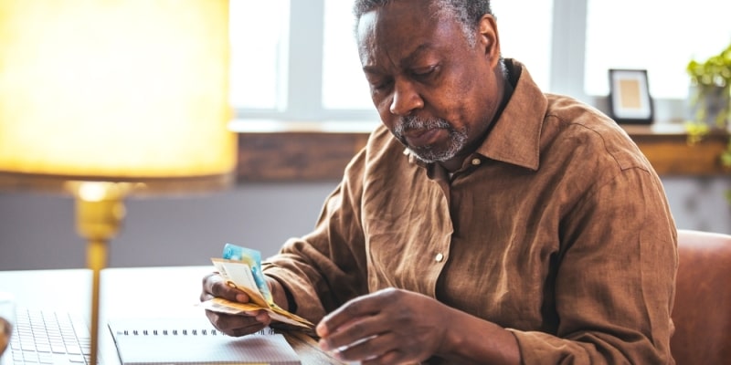 Um homem idoso negro com notas de dinheiro em uma das mãos. Ele está sentado em uma poltrona e na sua frente tem uma mesa, um caderno de anotações e um notebook. Imagem para ilustrar a matéria sobre superendividamento.