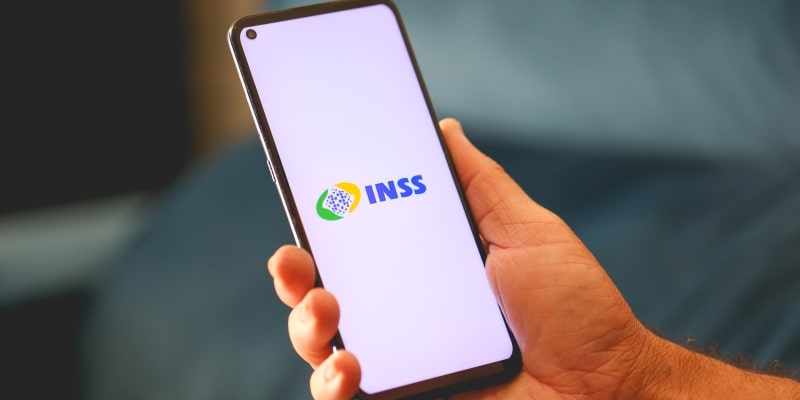 Um mão masculina segurando um celular com a logo do INSS. Imagem para ilustrar a matéria sobre corte de benefícios do INSS.