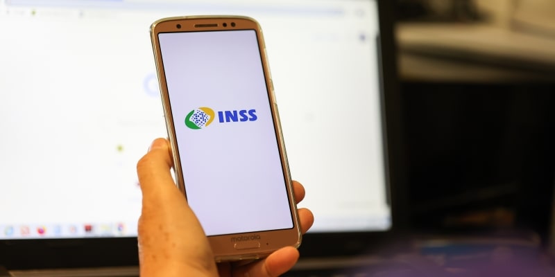 Uma mão idosa segurando um celular com a tela do INSS. Imagem para ilustrar a matéria sobre descontos associativos do INSS.