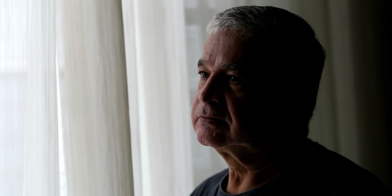 Um idosos triste, olhando para o horizonte, ao lado de uma janela com cortina fechada. Imagem para ilustrar a matéria sobre o número de denúncias de violência contra idosos.