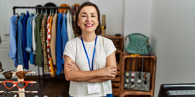 Uma mulher, em torno de 50 anos, de braços cruzados e sorrindo. Aos fundos, roupas e acessórios de sua loja. Imagem para ilustrar a matéria sobre qualquer profissão pode ser MEI.