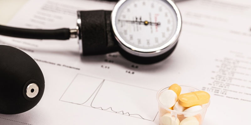 Medicação para hipertensão, um esfigmomanômetro e exames médicos em segundo plano. Imagem para ilustrar a matéria sobre 10 dúvidas frequentes sobre a hipertensão.