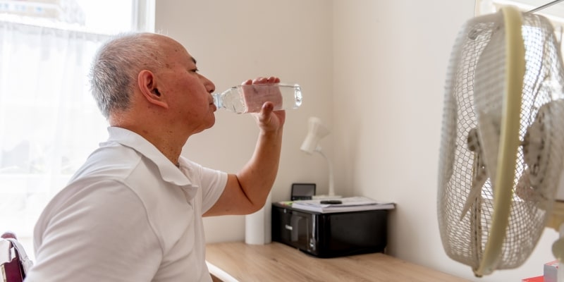 Um homem idosos bebendo água em um quarto, em frente a um ventilador. Imagem para ilustrar a matéria sobre calor extremo.