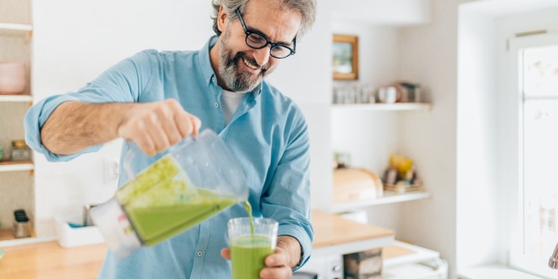 Um homem idoso sorrindo enquanto enche um copo com vitamina para complementar a alimentação e evitarr a sarcopenia.