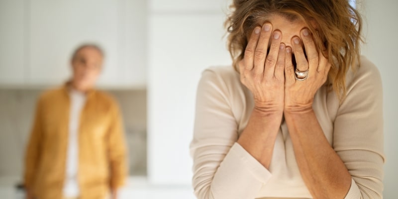 Um mulher que sofre de violência patrimonial triste, com as mãos no rosto, enquanto chora. Ao fundo, a imagem desfocada de um homem.