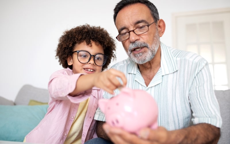 Avô e neto colocando dinheiro no cofrinho, sentados no sofá em uma sala de estar. Imagem para ilustrar a matéria sobre dicas para economizar dinheiro que realmente funcionam.