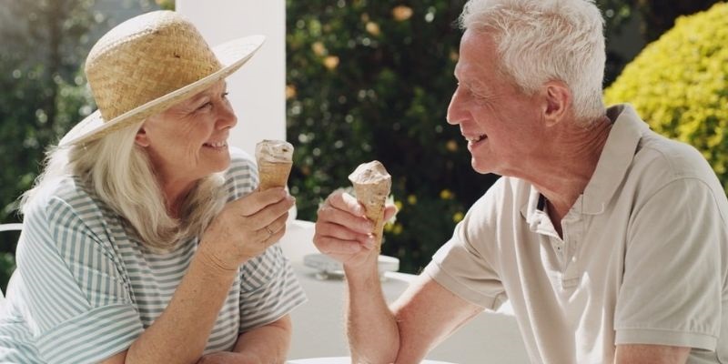 Casal caucasiano, em uma varanda, num dia quente de verão. Cada um segura um sorvete de casquinha, enquanto se olham e sorriem. Imagem para ilustrar a matéria sobre envelhecer com saúde.