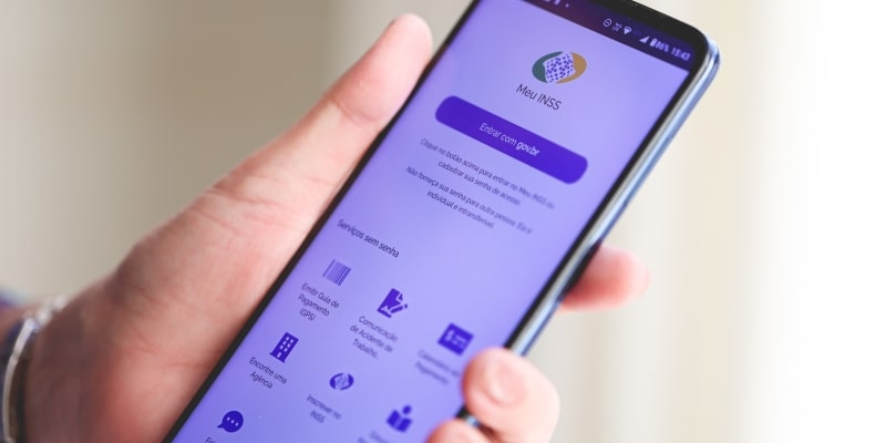 Uma mão segurando um celular, com o aplicativo do INSS aberto. Imagem para ilustrar a matéria sobre falhas no sistema do INSS.