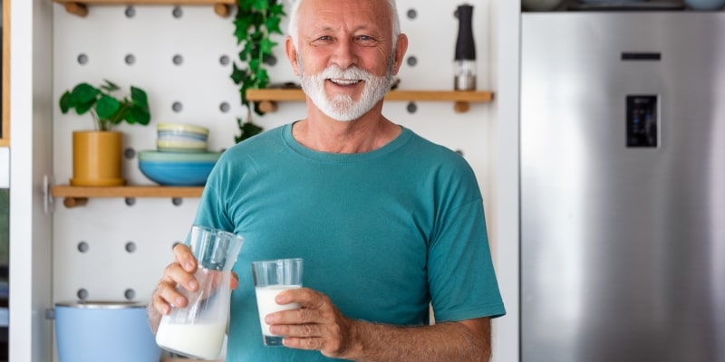 Um homem idoso sorrindo enquanto segura um copo e um jarra de leite, que também é fonte de proteínas.