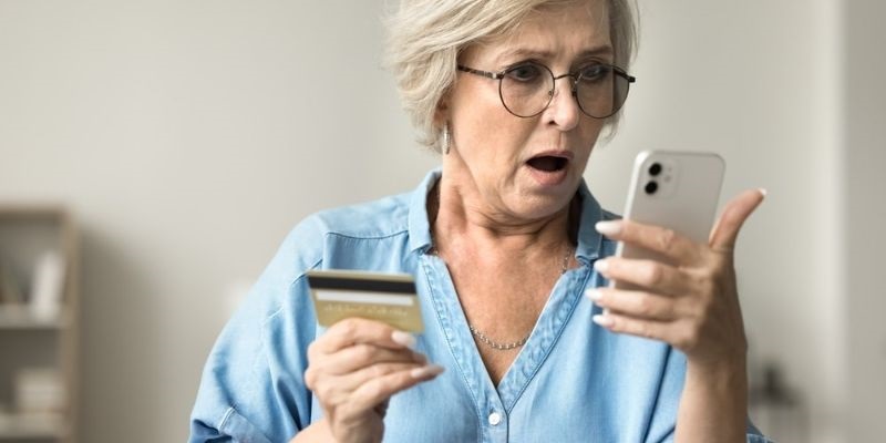 Uma mulher idosa segurando um cartão de crédito e olhando assustada para o celular. Imagem para ilustrar a matéria sobre apostas online.