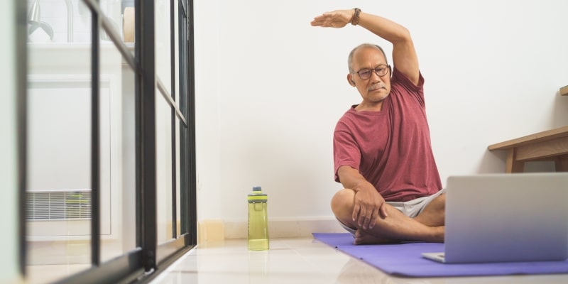 Um homem idoso treinando e esticando o braço, enquanto acompanha exercícios pelo laptop. Imagem para ilustrar a matéria sobre exercícios físicos beneficiam idosos com câncer.