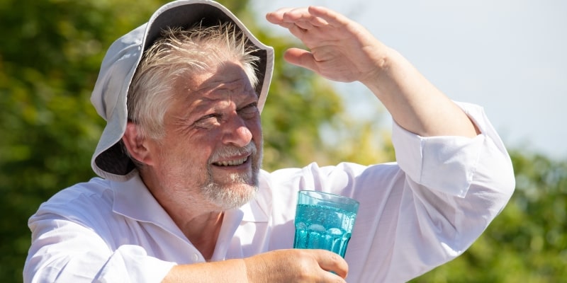 Um homem idoso com chapéu de sol tapando o rosto do sol. Ele bebe água de um copo. Imagem para ilustrar a matéria de mudanças climáticas.