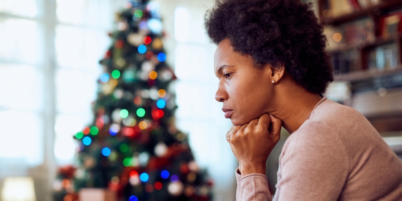 Uma mulher negra, apoiando o rosto nas mãos e lidando com a ansiedade no fim do ano. Ao fundo, uma árvore de natal. 