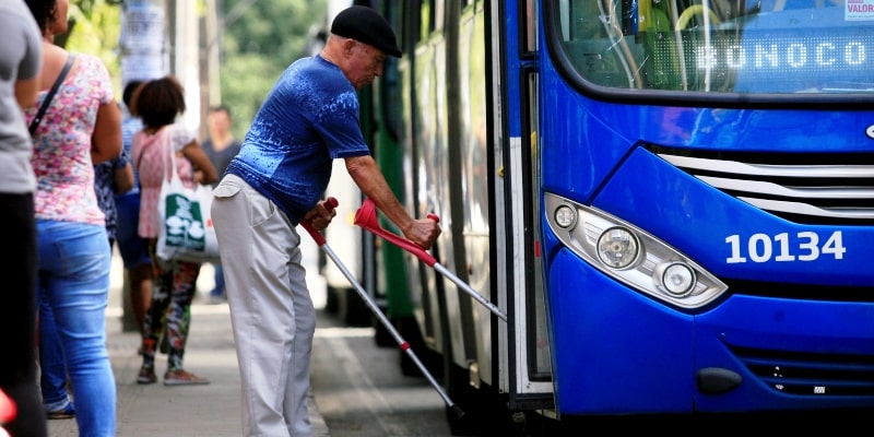 Um homem idoso de muletas tentando subir em um ônibus. Imagem para representar a matéria sobre longevidade com qualidade de vida.