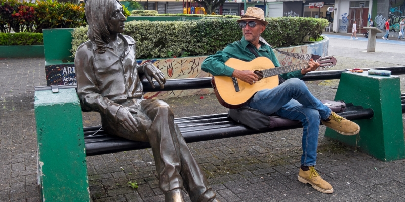 Um homem idoso tocando violão, sentado em um banco de praça, ao lado de uma estátua, em San José, capital da Costa Rica, um dos melhores países para viver bem gastando menos.