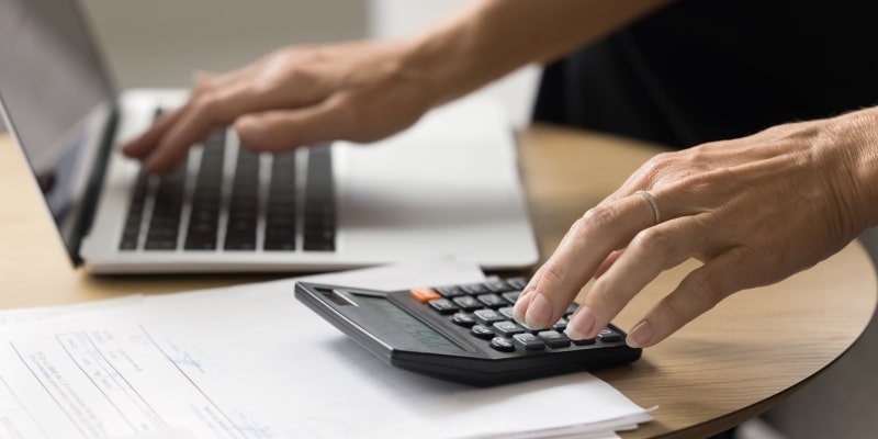 Mãos de uma pessoa em uma calculadora e em um computador, fazendo contas para decidir de vale a pena pagar as despesas de início de ano à vista.