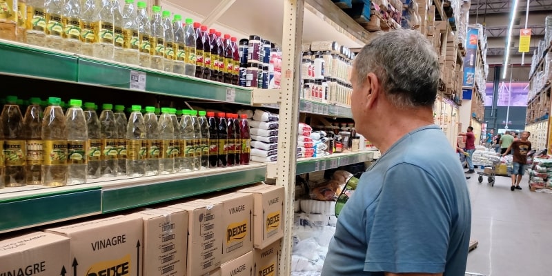 Um homem idoso em um supermercado olhando a prateleira de vinagre. Imagem para ilustrar a matéria sobre a alta dos preços da comida.