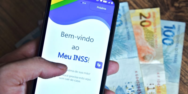 O aplicativo chamou Meu INSS na tela de um celular que um homem está segurando e notas de dinheiro brasileiro ao fundo. Imagem para ilustrar a matéria sobre os descontos indevidos na aposentadoria.