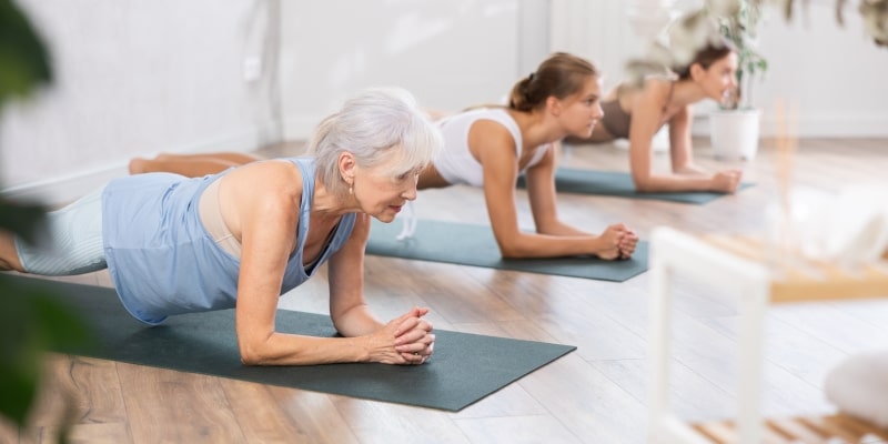 Uma mulher idosa fazendo prancha, como um exercício funcional de sua prática de hábitos de vida saudável. Ela está ao lado de duas mulheres jovens.