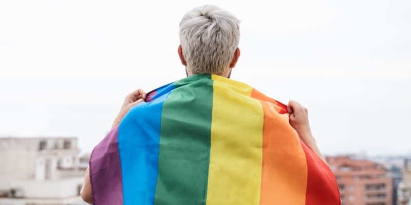 Um homem de cabelos brancos com a bandeira LGBT nas costas, representando a matéria sobre longevidade com orgulho.