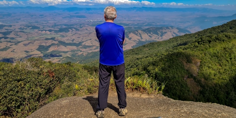 Um homem idoso contemplando a vista de um vale após subir um morro em uma trilha. Imagem para ilustrar a matéria sobre movimento.