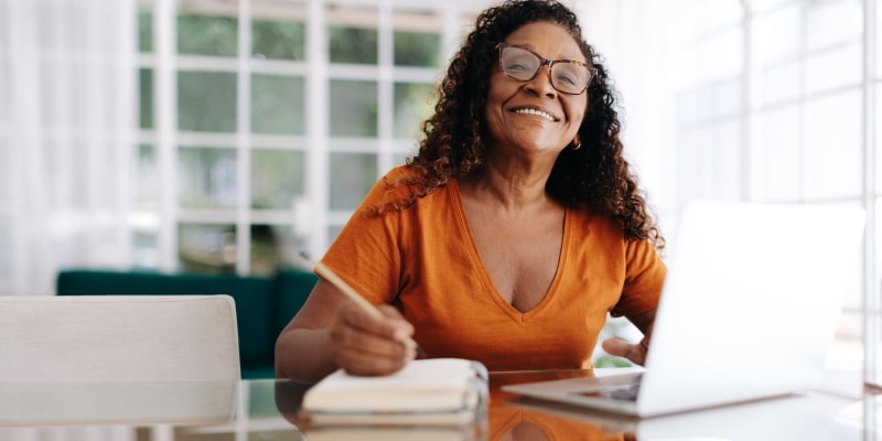 Uma mulher idosa e negra sorrindo enquanto mexe no computador e faz anotações. Imagem para ilustrar a matéria sobre o cérebro da mulher madura.