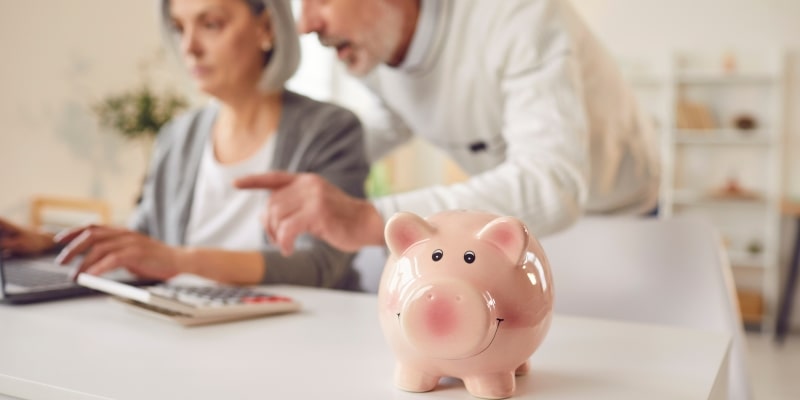 Um cofre de porquinho em destaque. Ao fundo, um casal conversando sobre finanças em frente a um computador e ao lado de uma calculadora. Imagem para ilustrar a matéria sobre o score de crédito dos idosos.