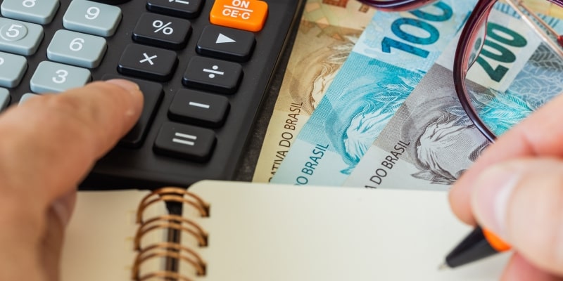 Uma mão segurando uma caneta para fazer anotações em um caderno. Também há notas de dinheiro, uma calculadora e um óculos. Imagem para ilustrar organização financeira nos cuidados com o décimo terceiro.