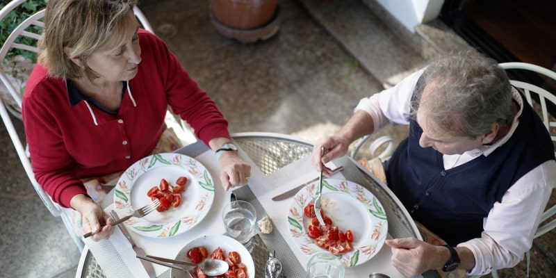 Casal de idosos desfrutando de uma refeição leve ao ar livre. A vista é de cima para baixo e eles comem salada de tomate. Imagem para ilustrar a matéria sobre o sistema imunológico resistente.
