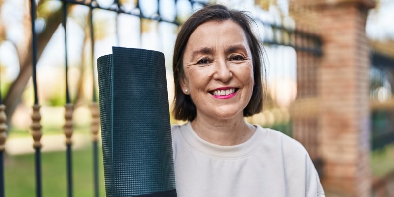 Mulher madura sorrindo para a câmera e segurando um tapete de ioga que está enrolado, em um parque. Imagem para ilustrar a matéria sobre 150 minutos de atividade física.