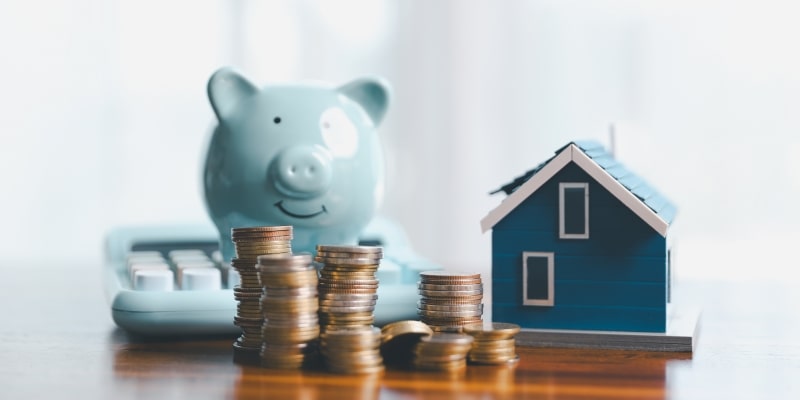 Moedas empilhadas em destaque. Ao fundo, um cofre de porquinho, uma miniatura de casa e uma calculadora. Imagem para ilustrar a matéria sobre benefícios não resgatados após falecimento.