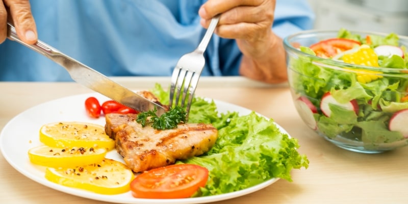 Uma pessoa cortando um pedaço de carne em um prato com salada. Imagem para ilustrar a matéria sobre controlar o colesterol.