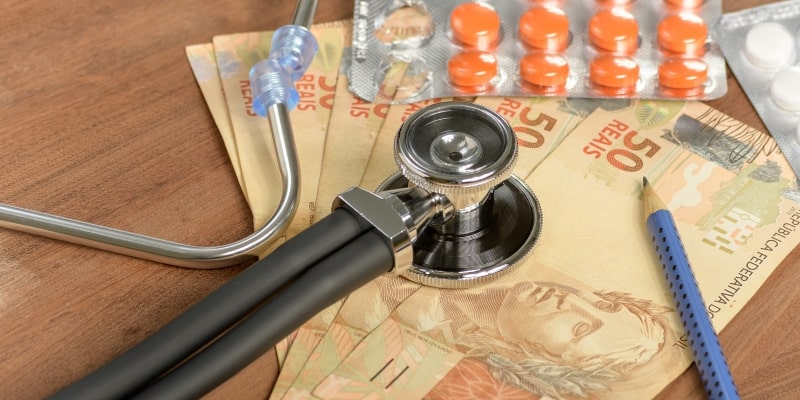 Estetoscópio médico sobre dinheiro brasileiro, um lápis e pacotes de remédios sobre fundo de madeira. Imagem para ilustrar a matéria sobre o preço dos planos de saúde.