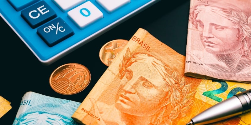 Notas de dinheiro, moedas, uma caneta e uma calculadora, ilustrando a matéria sobre a restituição do imposto de renda 2025.