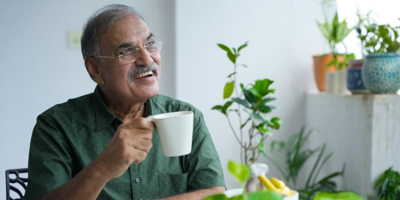 Um senhor idoso, sentando em frente a uma mesa, sorrindo enquanto segura uma xícara de chá. Imagem para ilustrar a matéria sobre ser positivo.