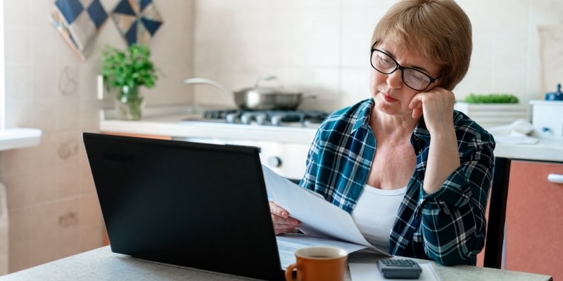 Uma mulher com mais de 50 anos, em sua cozinha, mexendo no computador e olhando papéis de demissão, desanimada. Imagem para ilustrar a matéria sobre blindar as finanças.