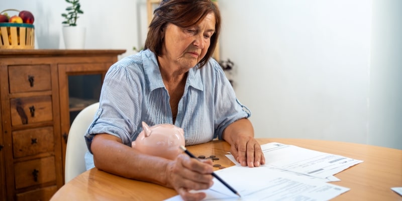 Mulher sênior sentada à mesa revisando notas financeiras com moedas e cofrinho, refletindo sobre a gestão de despesas e desafios financeiros em casa. Imagem para ilustrar a matéria sobre custos com moradia.