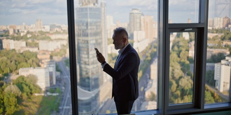 Um homem idoso, em um escritório de janela panorâmica de vidro, chocado do ao olhar uma mensagem no celular. Imagem para ilustrar a matéria sobre demissão por idade.