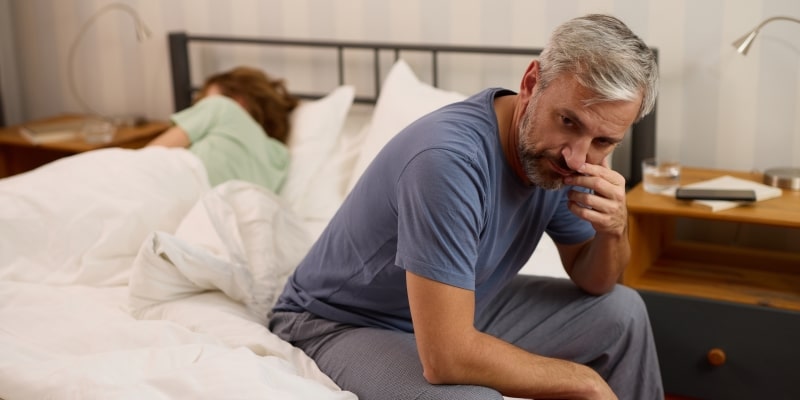 Um home com disfunção erétil triste, sentado na cama. Ao seu lado, deitada na cama, está uma mulher, de costas.