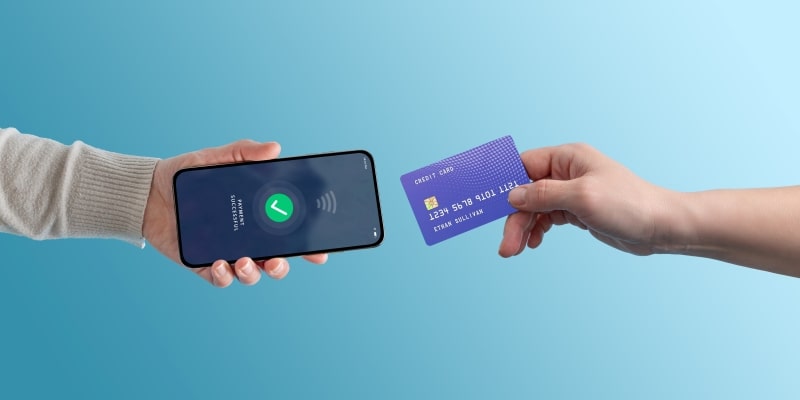 Mão de uma pessoa segurando um cartão de crédito enquanto o aproxima de um celular, na mão de outra pessoa, para realizar o pagamento, ilustrando a matéria sobre golpe do toque fantasma.